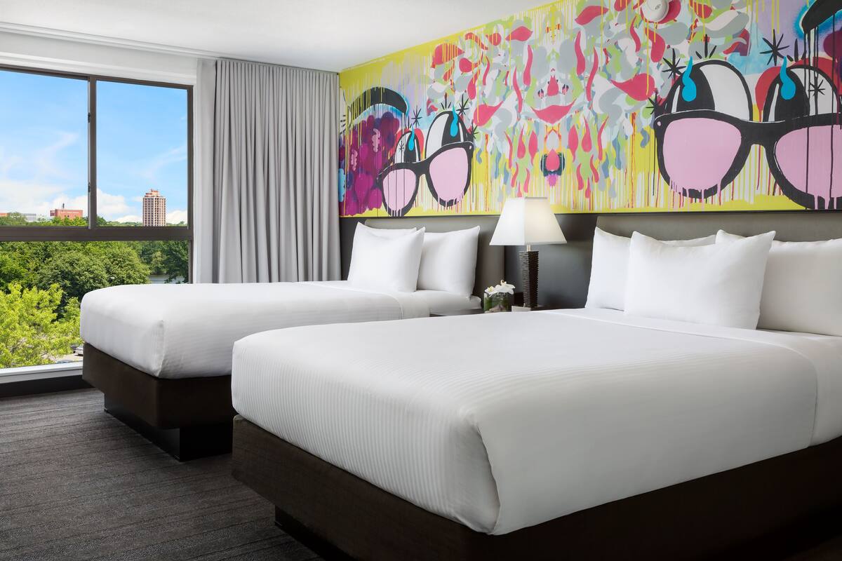 Une chambre d’hôtel avec deux lits, une grande fenêtre et un tableau abstrait aux couleurs vives accroché au mur.