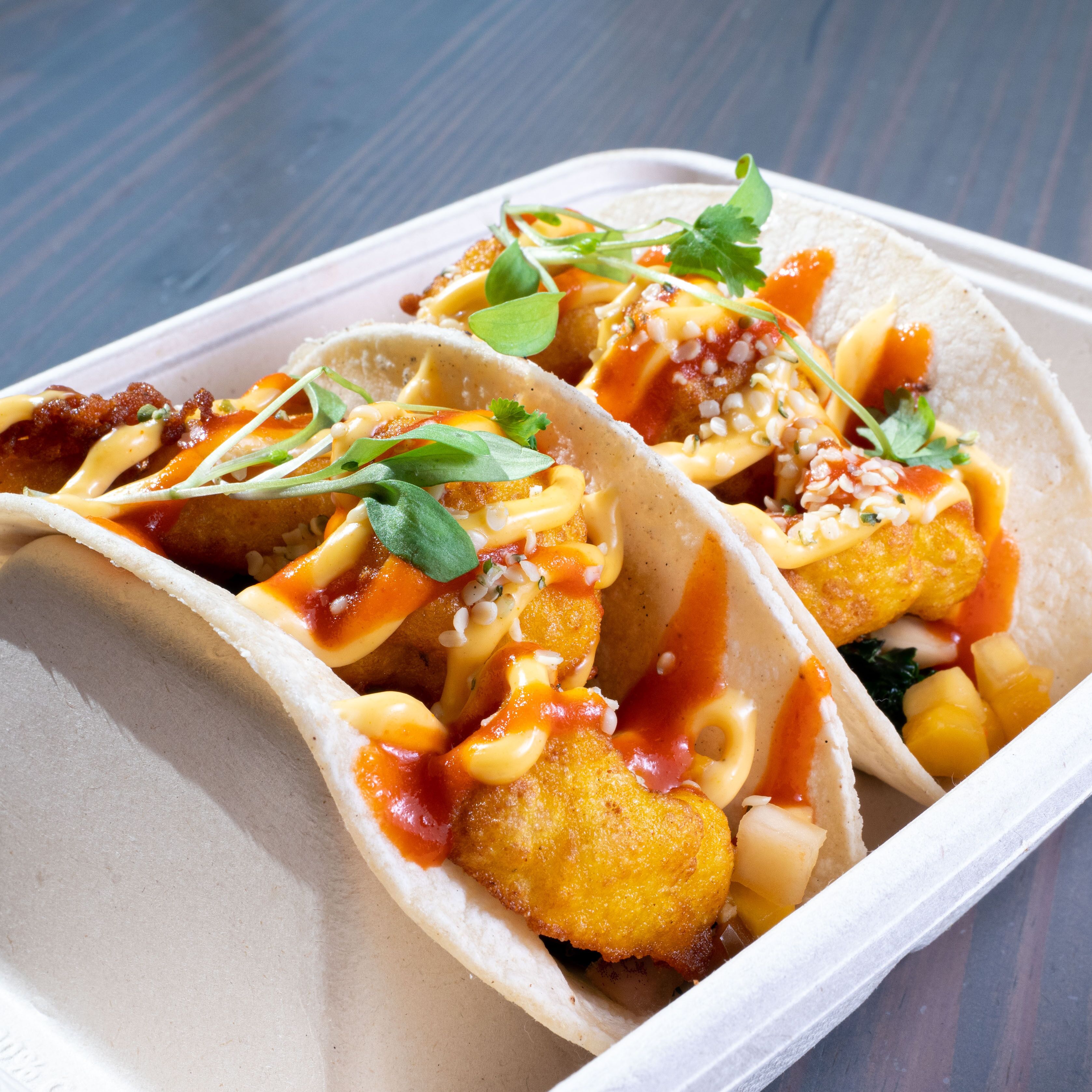 Two tacos with a crispy coating, garnished with fresh herbs and a drizzle of sauce, served in a white paper tray.
