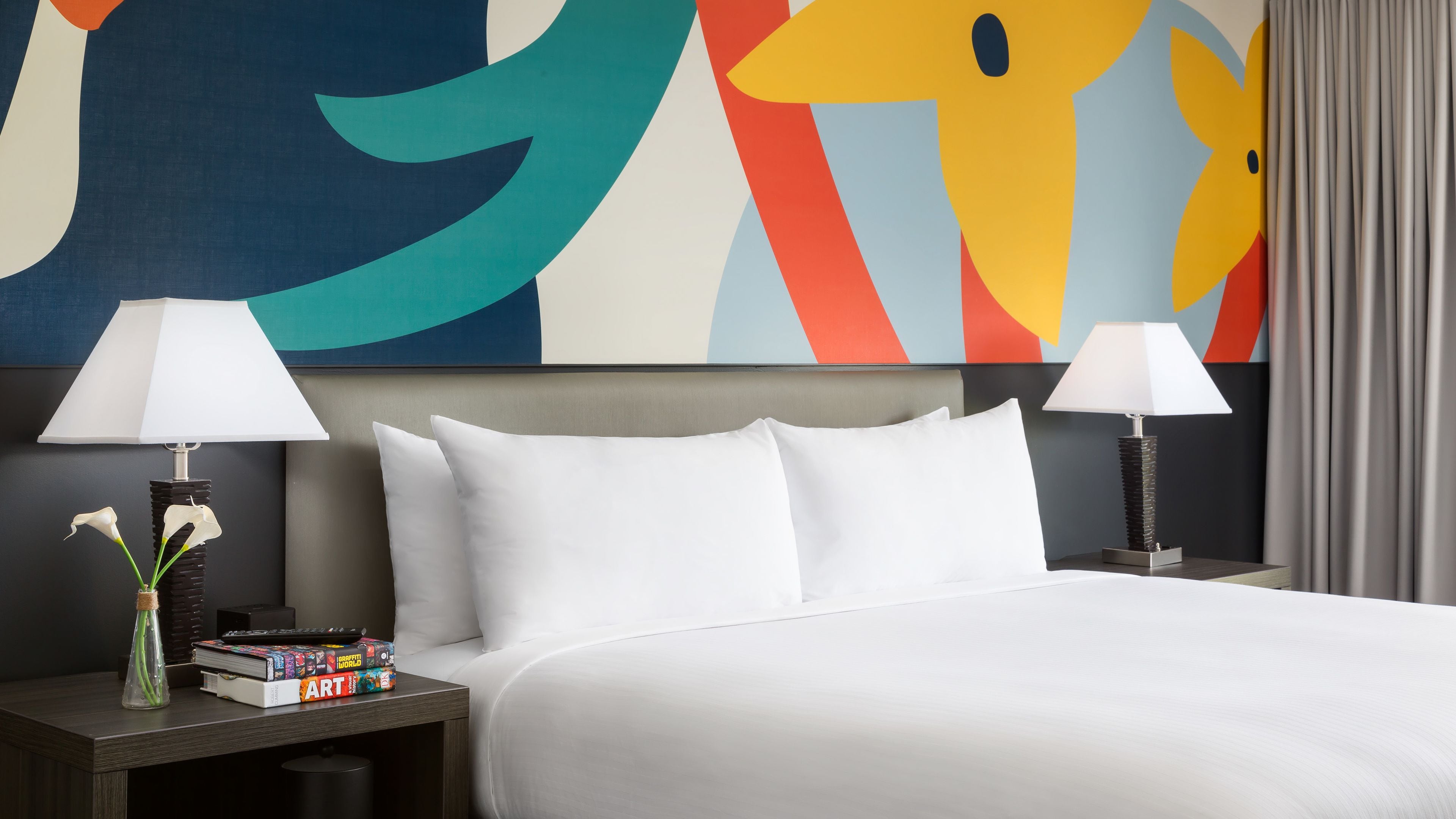 A hotel room with a large bed, two bedside tables with lamps, a colorful abstract painting on the wall, and a vase with flowers on one table.