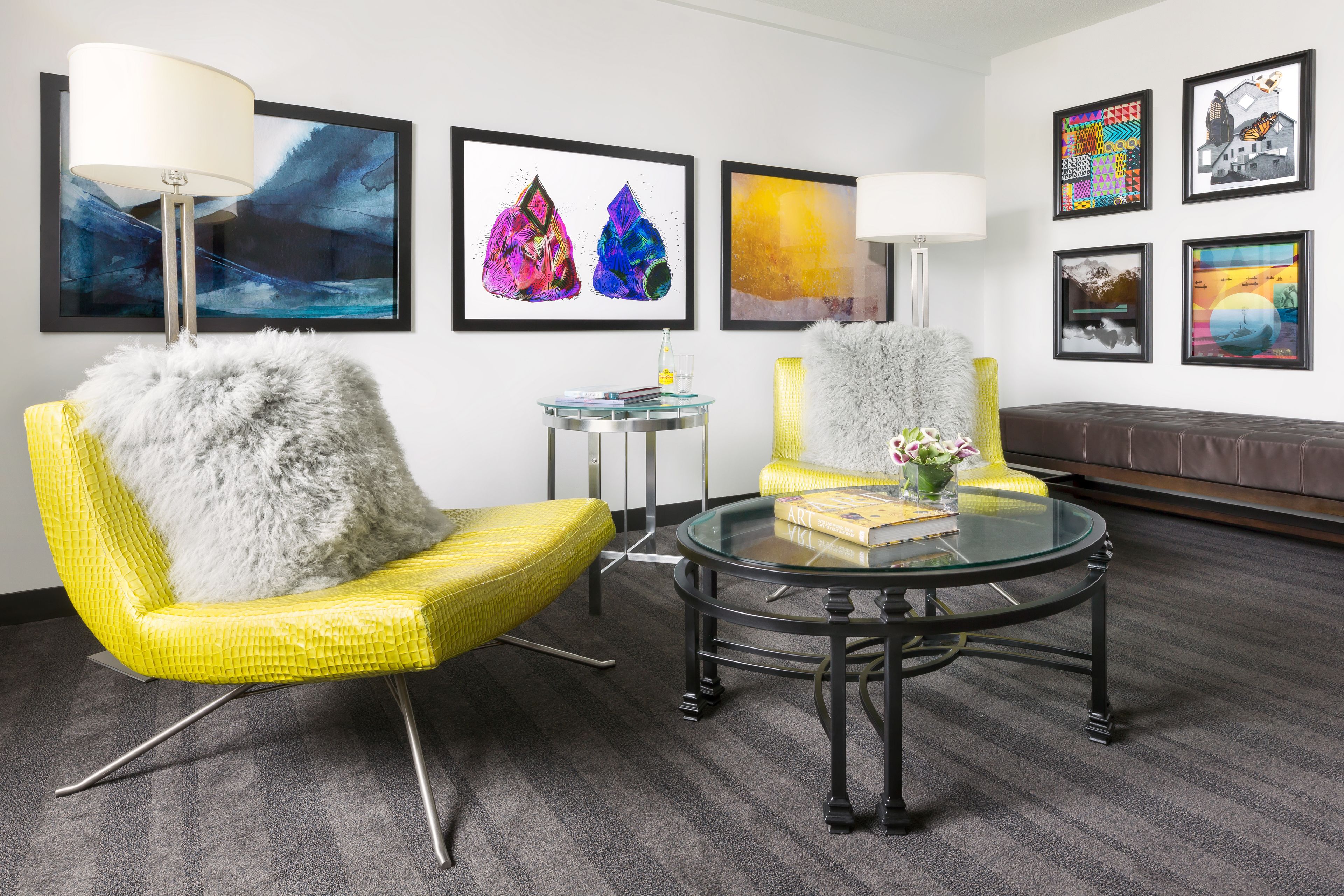 A modern room with a glass coffee table, two yellow chairs with fluffy cushions, a round side table, and a sofa.