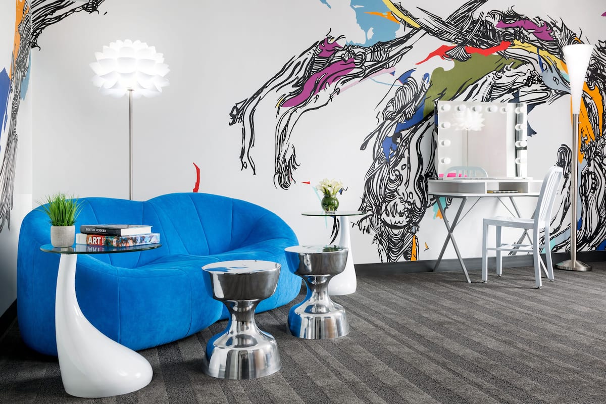 Une pièce moderne avec un canapé bleu, un lampadaire blanc, une coiffeuse avec un miroir et un mur orné d’œuvres d’art abstraites.