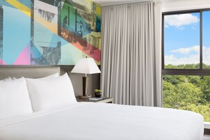 Hotelkamer met een groot bed, nachtkastje met lamp, een potplant en uitzicht op groen door een raam.