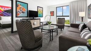 Una habitación de hotel moderna con un sofá, un sillón y una mesa de centro de vidrio. Hay un televisor de pantalla plana, una planta en maceta y obras de arte en las paredes.