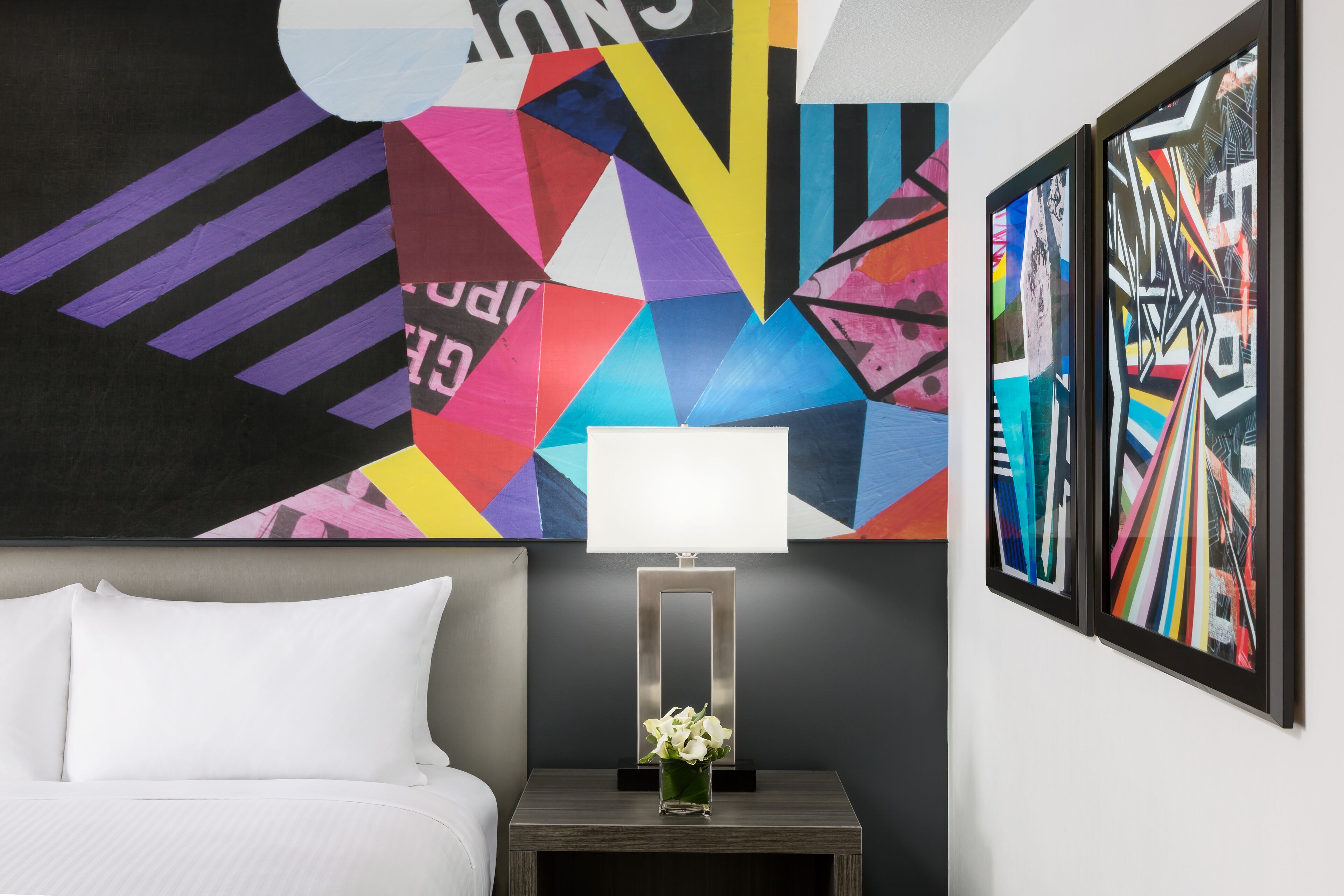 A modern hotel room with a bed, a nightstand with a lamp, and a colorful abstract painting on the wall.