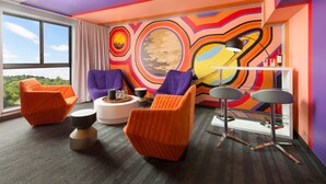 Una habitación moderna con sillas anaranjadas y moradas, una mesa redonda blanca y un mural abstracto lleno de color en la pared.