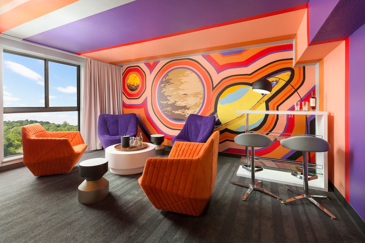 Une pièce moderne avec des chaises orange et violettes, une table ronde blanche et une fresque murale abstraite et colorée.