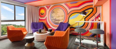 A modern room with orange and purple chairs, a white round table, and a colorful abstract mural on the wall.