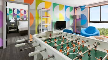 Una habitación con mesa de fútbol, televisor, estantería y ventana con vistas a zonas verdes.