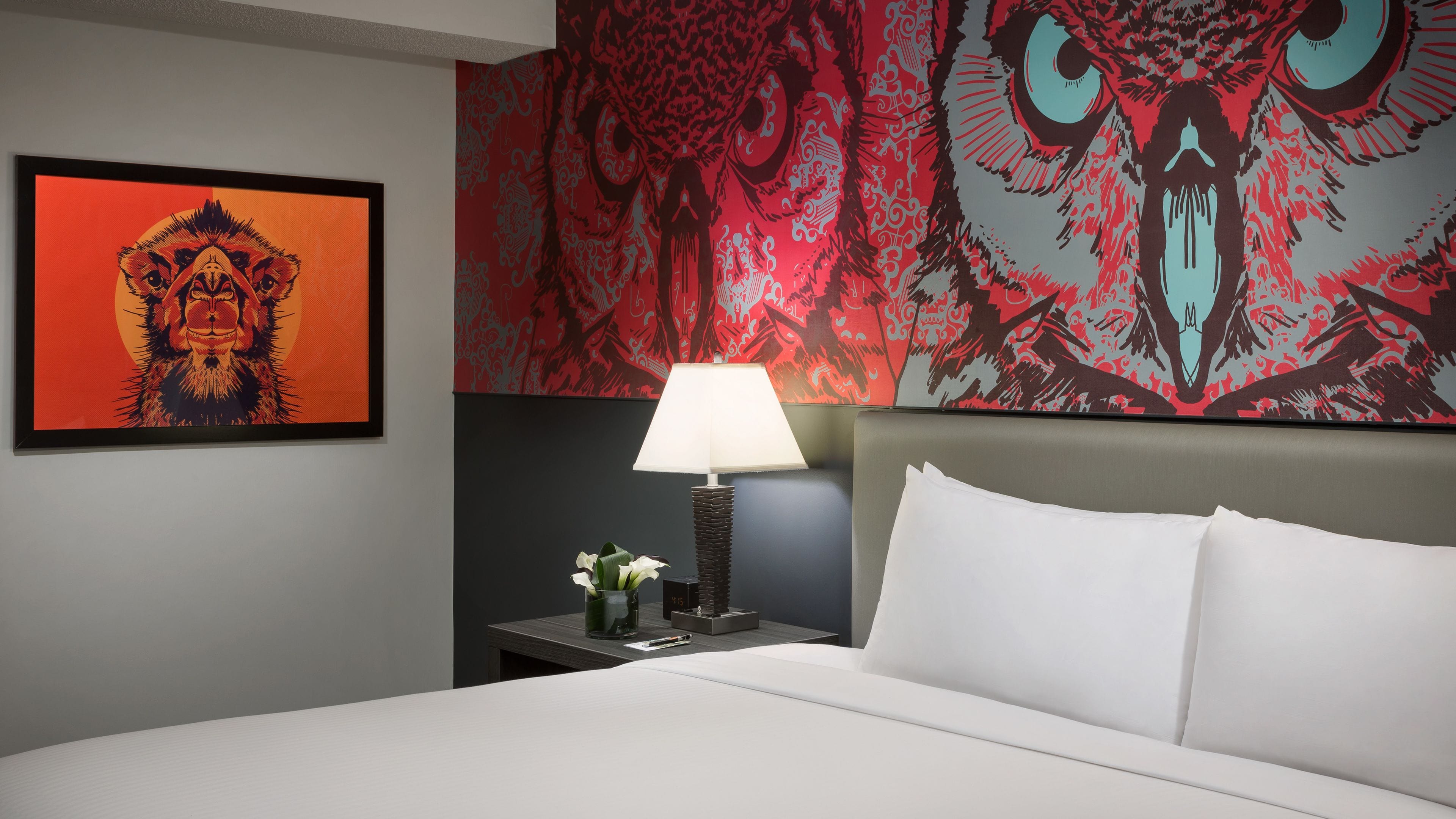 A hotel room with a bed, a nightstand with a lamp, a vase with flowers, and a large abstract painting on the wall.