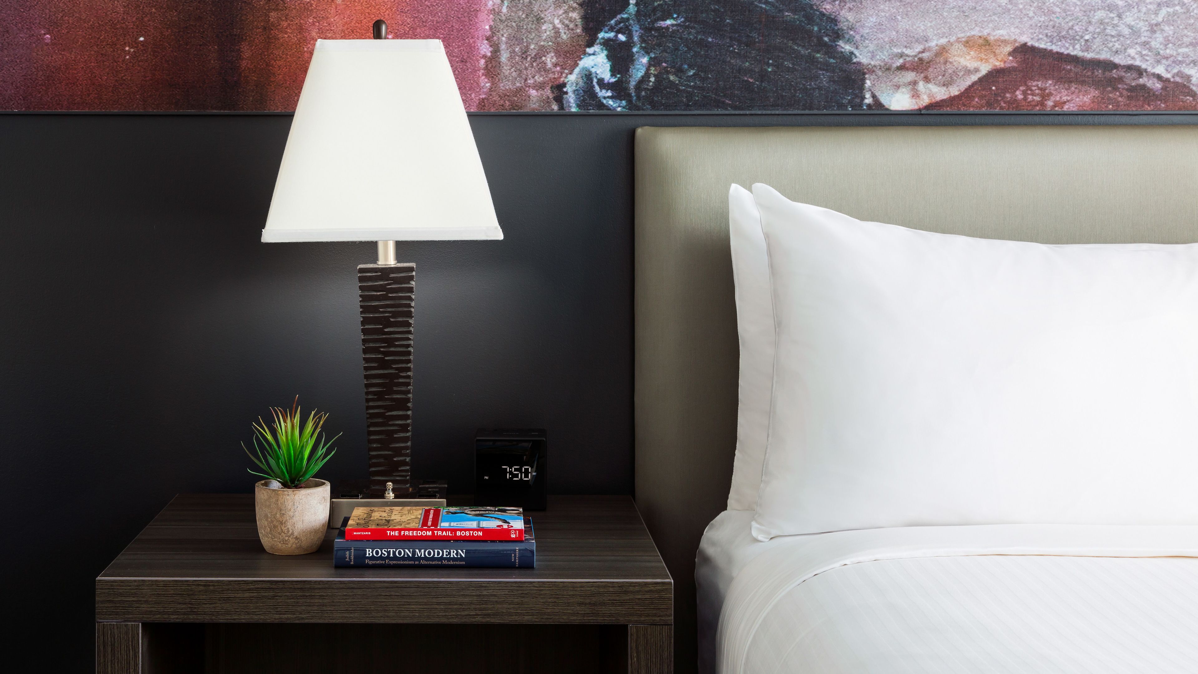 A neatly made bed with a white pillow and a headboard, a bedside table with a lamp, a small potted plant, and a few books.