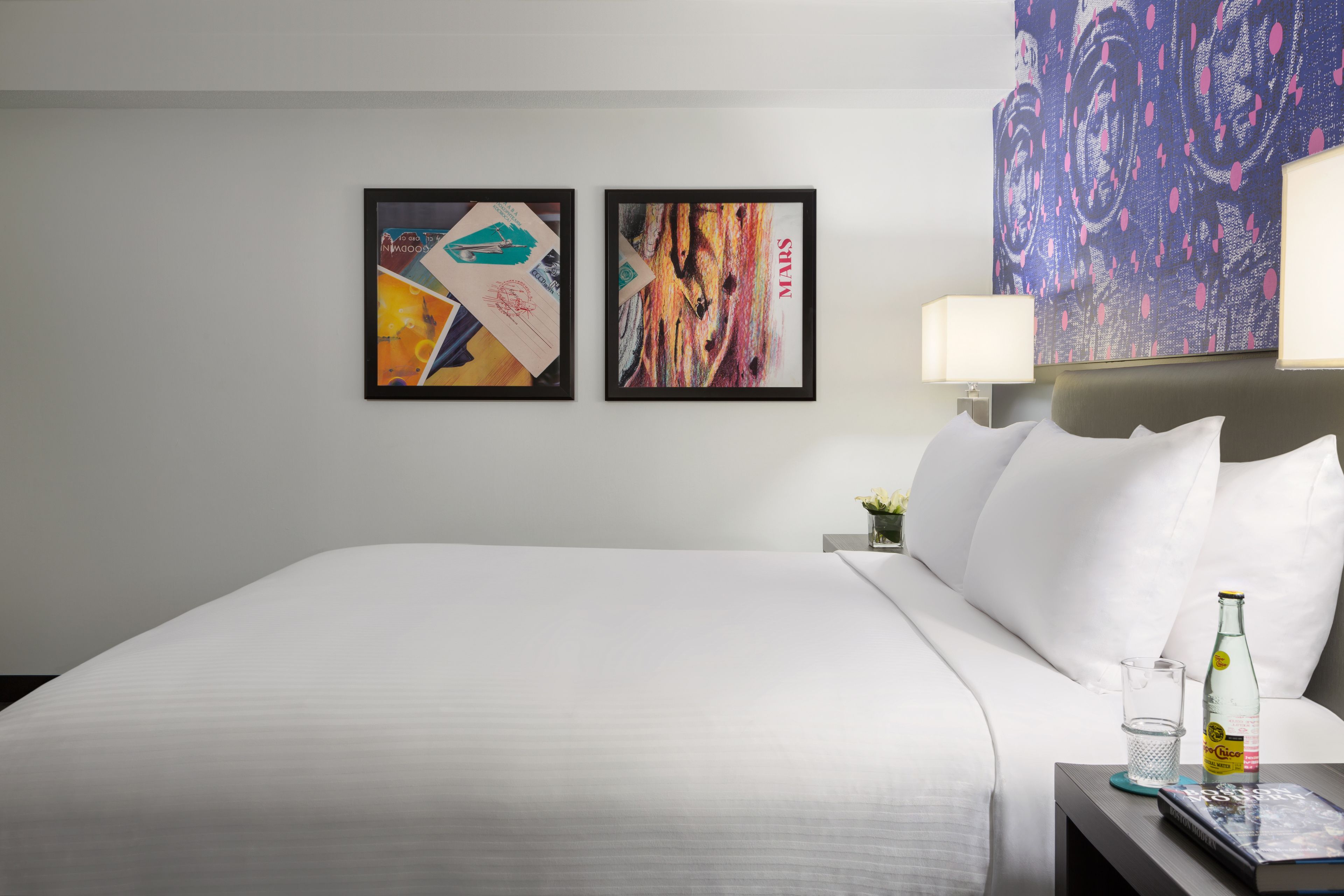 Habitación de hotel con una cama grande, dos cuadros enmarcados en la pared, una mesita de noche con una lámpara, una botella y un vaso.