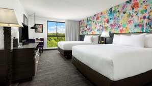 Habitación de hotel con dos camas, un escritorio, una silla, un televisor y un ventanal con vistas a zonas verdes.