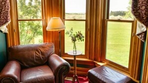 A room with a leather armchair, a small table with a lamp, and a view of a green landscape through large windows. - Country Hermitage B&B (Williamsburg)