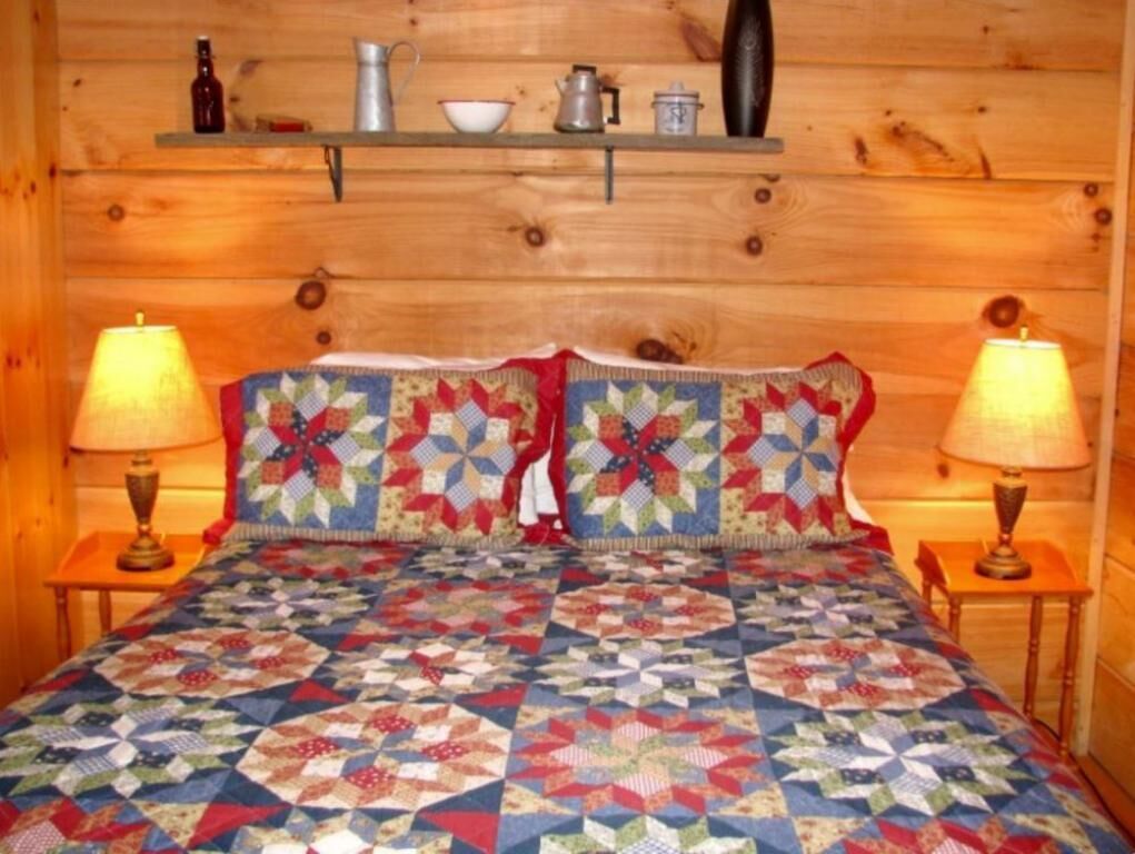 Interior de una cabaña de madera con una cama, dos lámparas y una estantería con diversos objetos.