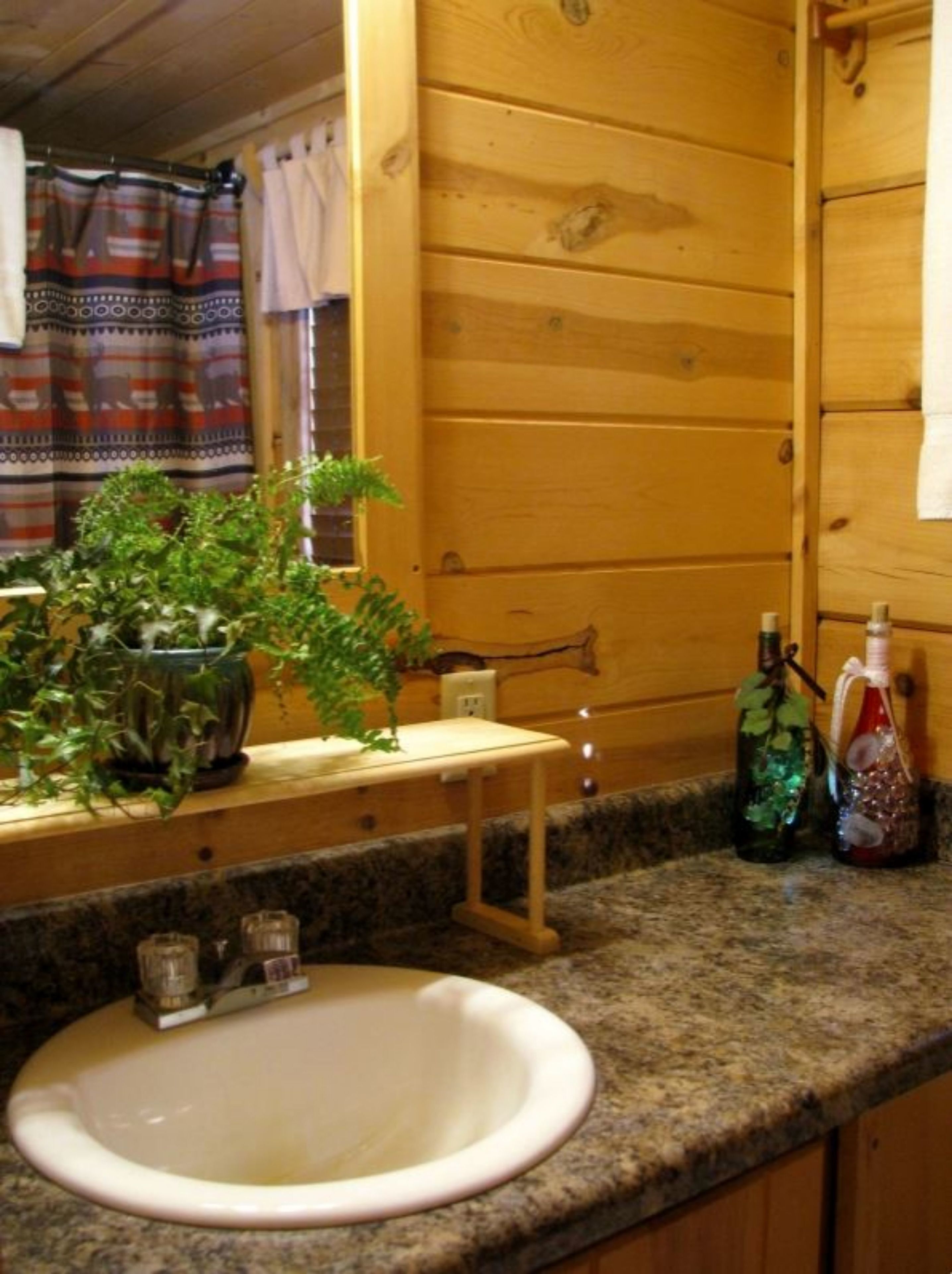 A wooden cabin bathroom with a granite countertop, a white sink, a mirror, a towel rack, and a window with curtains.
