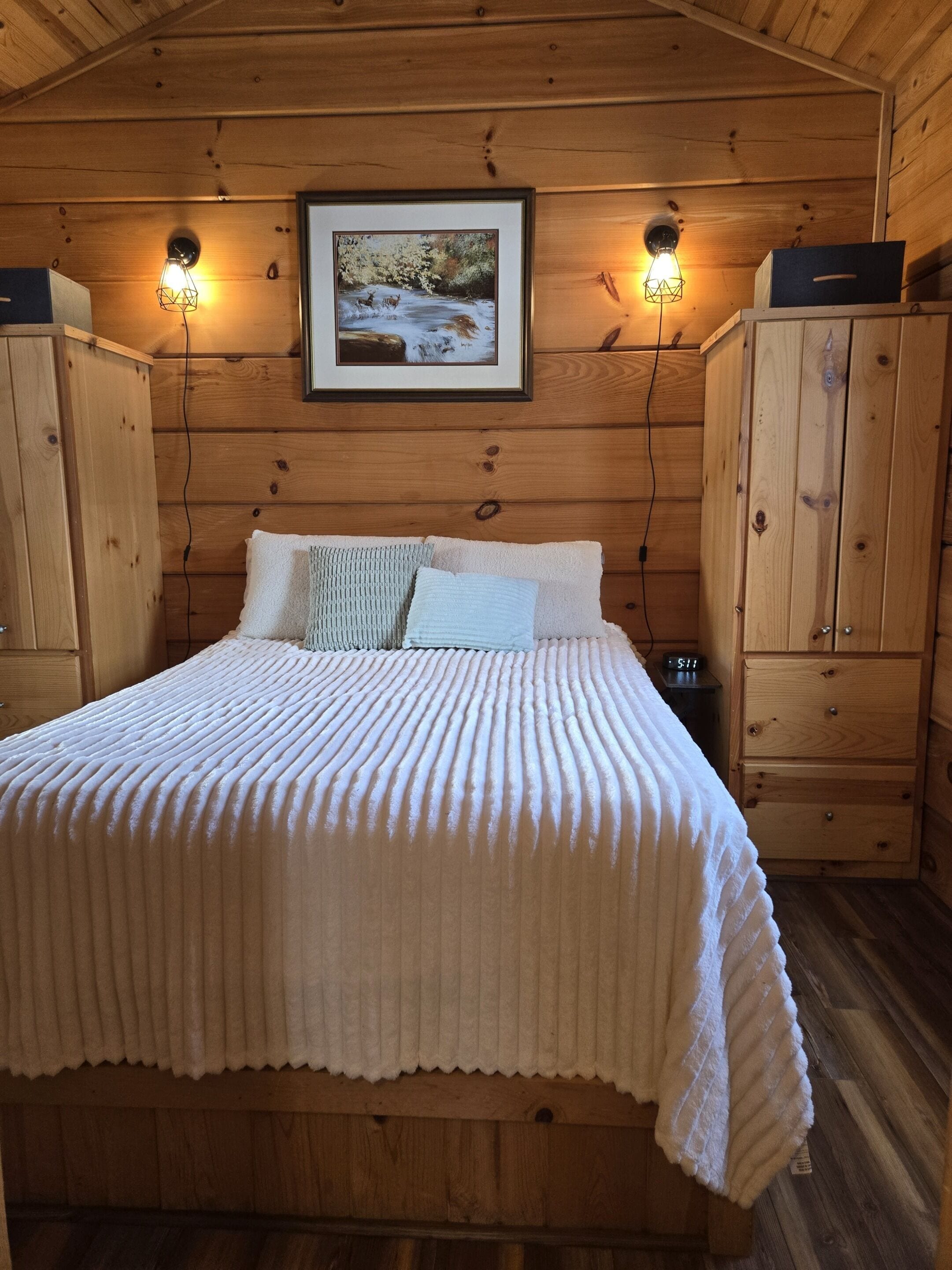 A wooden cabin bedroom with a bed, two wall-mounted lights, a framed picture, and wooden cabinets.