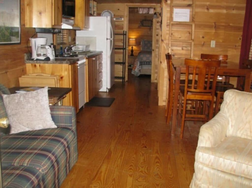 A wooden cabin interior with a kitchen, dining area, and a living room with a couch and chairs.
