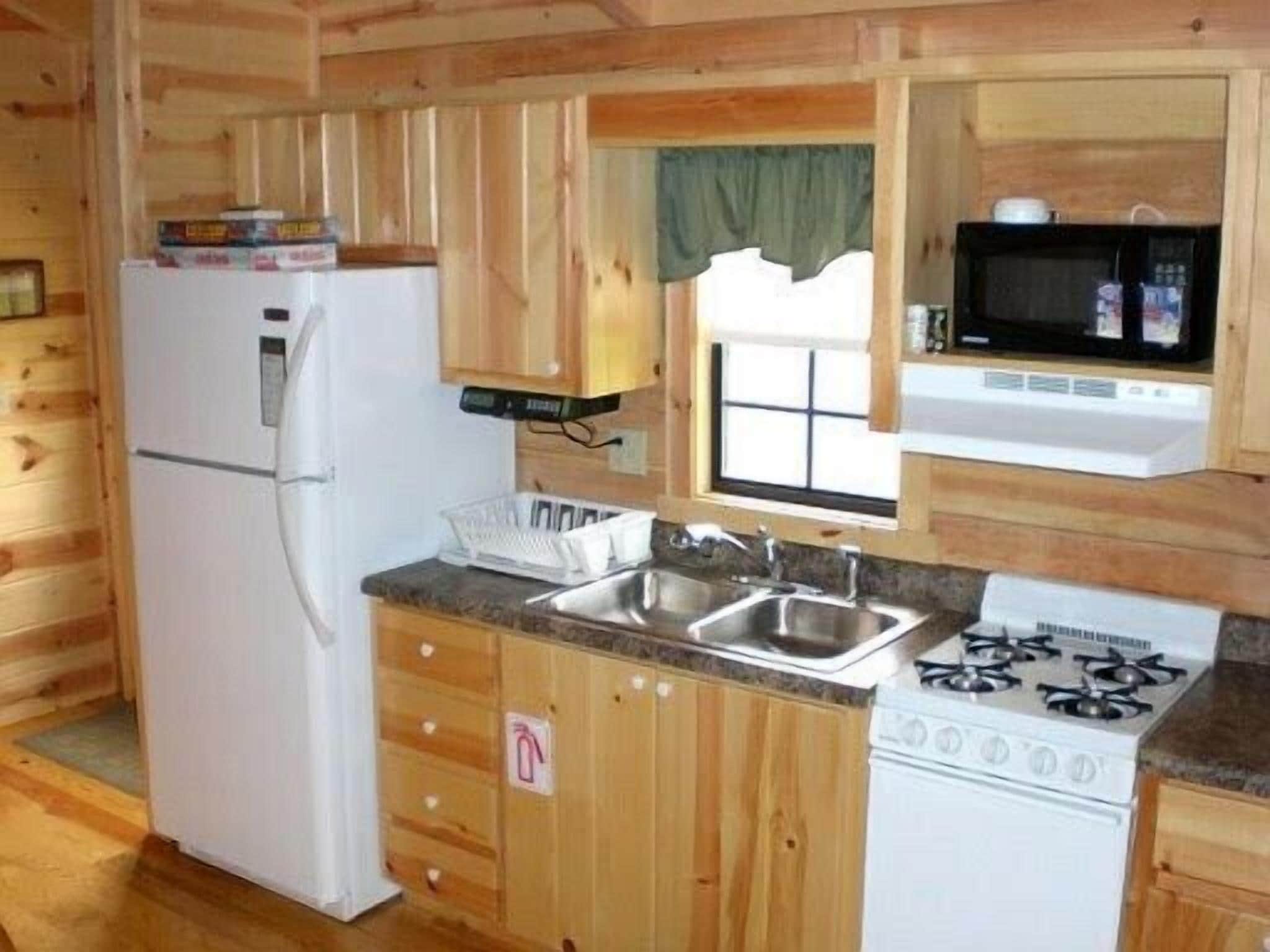 Une cuisine de taille réduite avec des murs en bois, un réfrigérateur, un four à micro-ondes, une cuisinière et une fenêtre ornée d’un rideau vert.