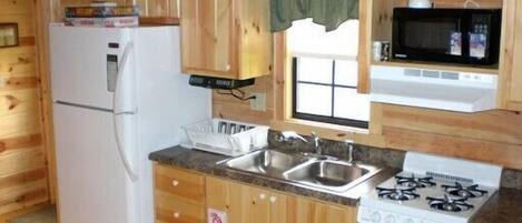 Une cuisine de taille réduite avec des murs en bois, un réfrigérateur, un four à micro-ondes, une cuisinière et une fenêtre ornée d’un rideau vert.