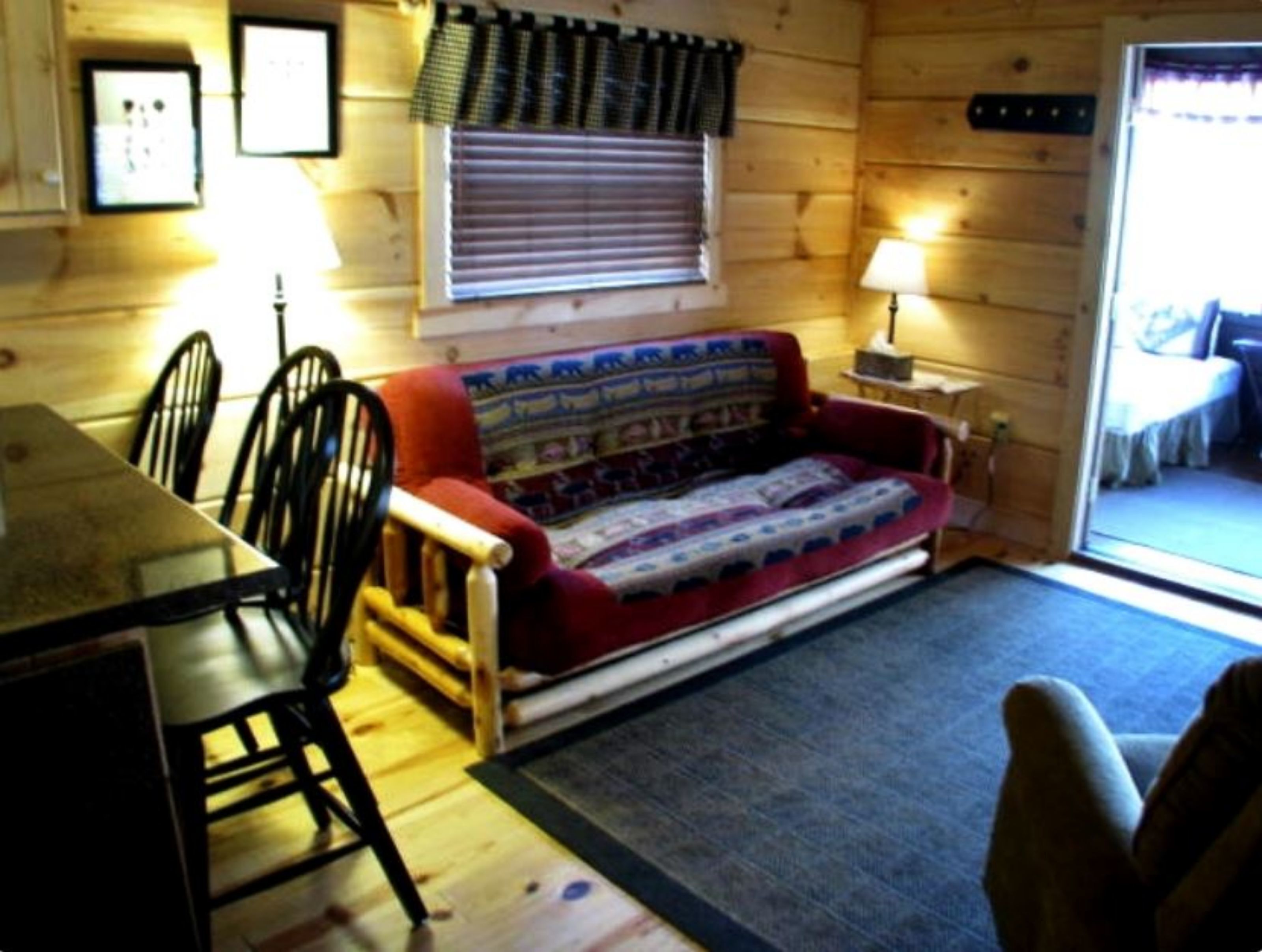 Ein gemütliches Blockhauss-Interieur mit einem Holzesstisch, Stühlen, einer Couch und einem Fenster mit Jalousien.