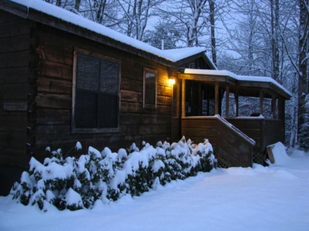 Eine Holzblockhütte mit Veranda und einer beleuchteten Außenlampe, umgeben von verschneitem Gelände und Bäumen.