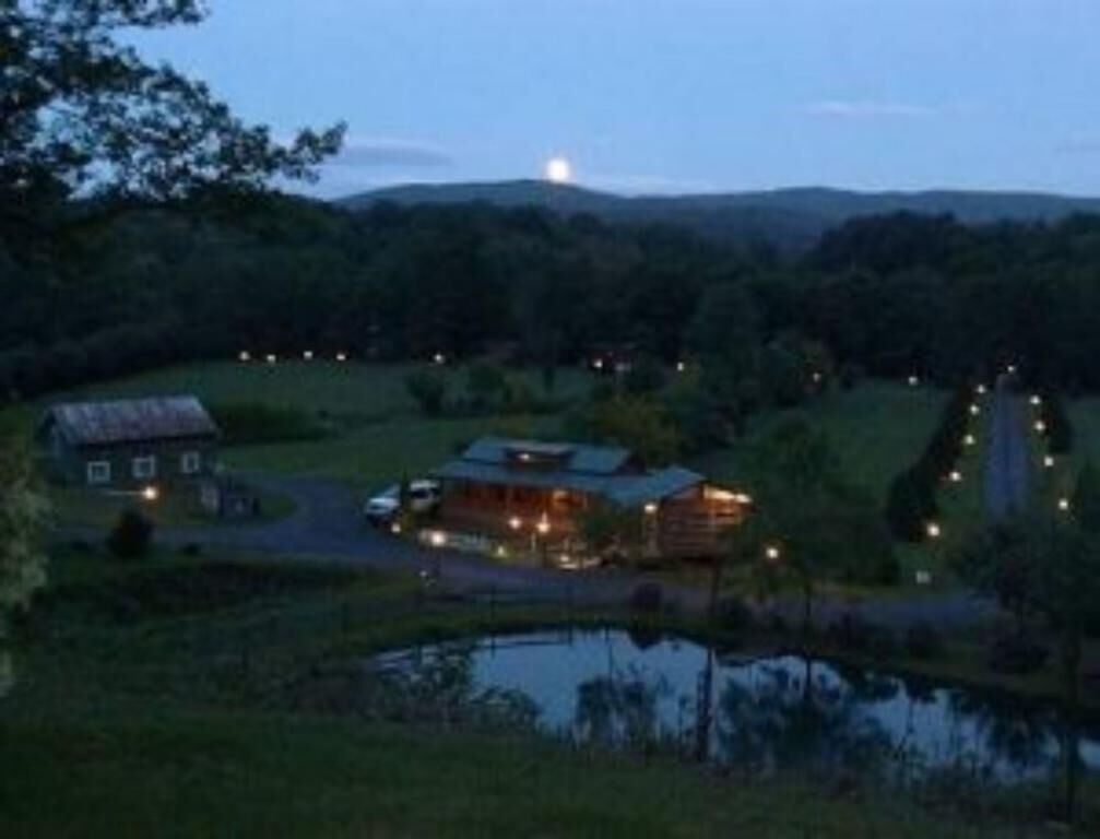 A rustic cabin with a pond in front, surrounded by greenery and hills.