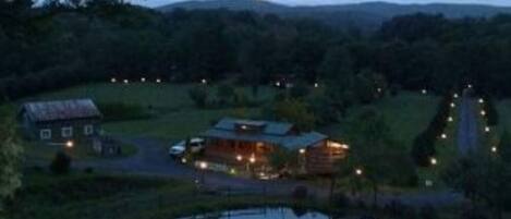 A rustic cabin with a pond in front, surrounded by greenery and hills.