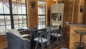 A wooden cabin interior with a dining table, chairs, a sofa, a patterned rug, and a kitchen area with a stove and cabinets. - Linville River Log Cabins (Newland)