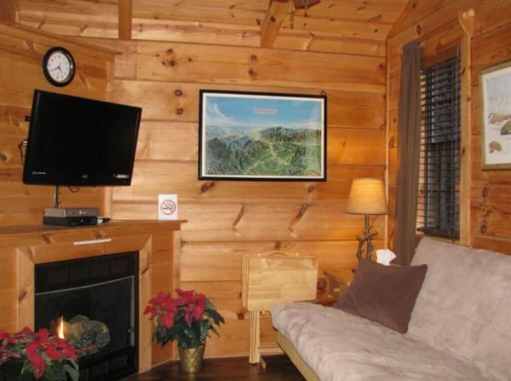 A cozy cabin interior with a fireplace, a television mounted on the wall, a framed landscape picture, and a couch with a pillow.