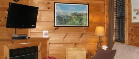 A cozy cabin interior with a fireplace, a television mounted on the wall, a framed landscape picture, and a couch with a pillow.