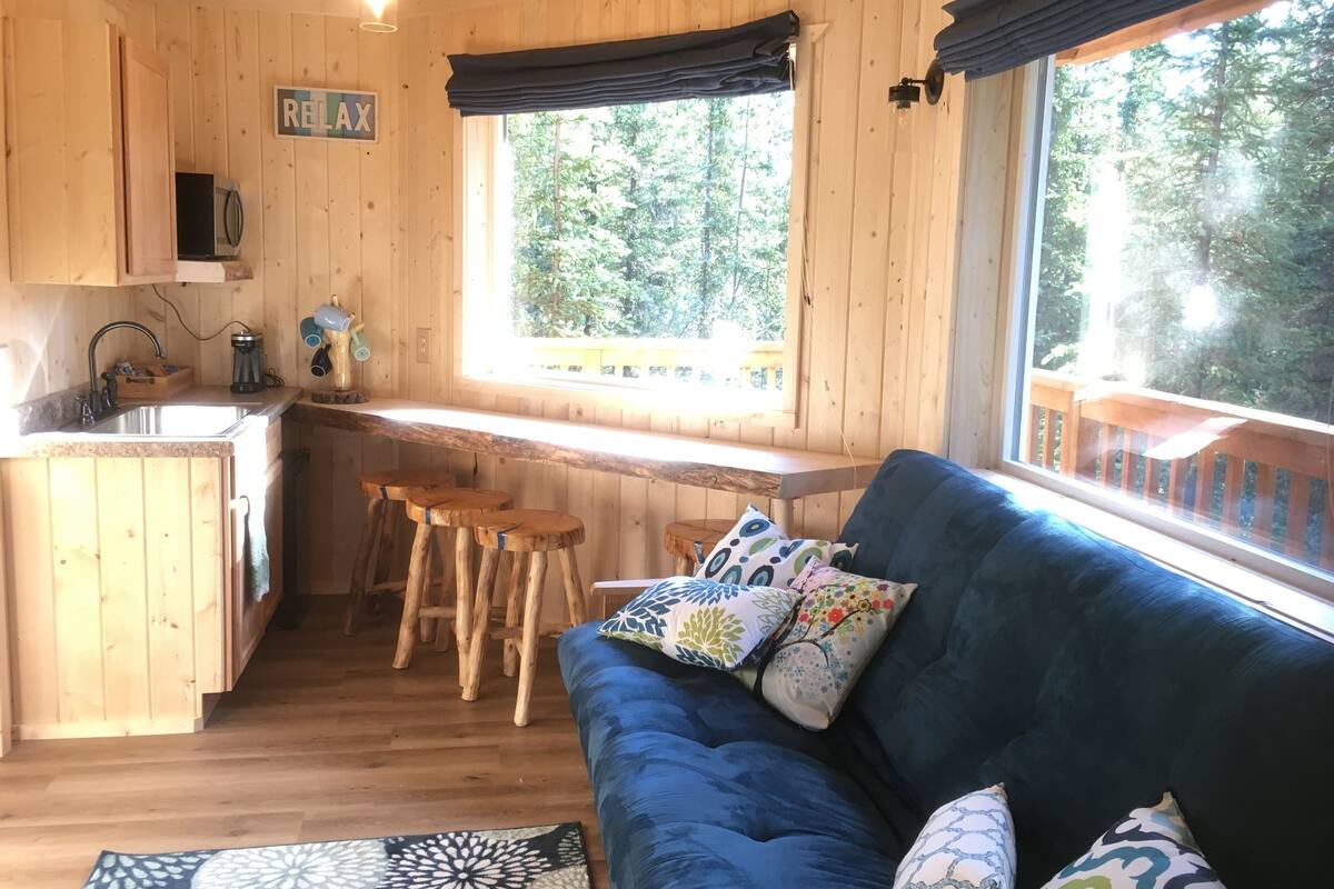 Interior de una cabaña con kitchenette, un sofá azul con cojines estampados y una ventana con vistas a una terraza de madera.