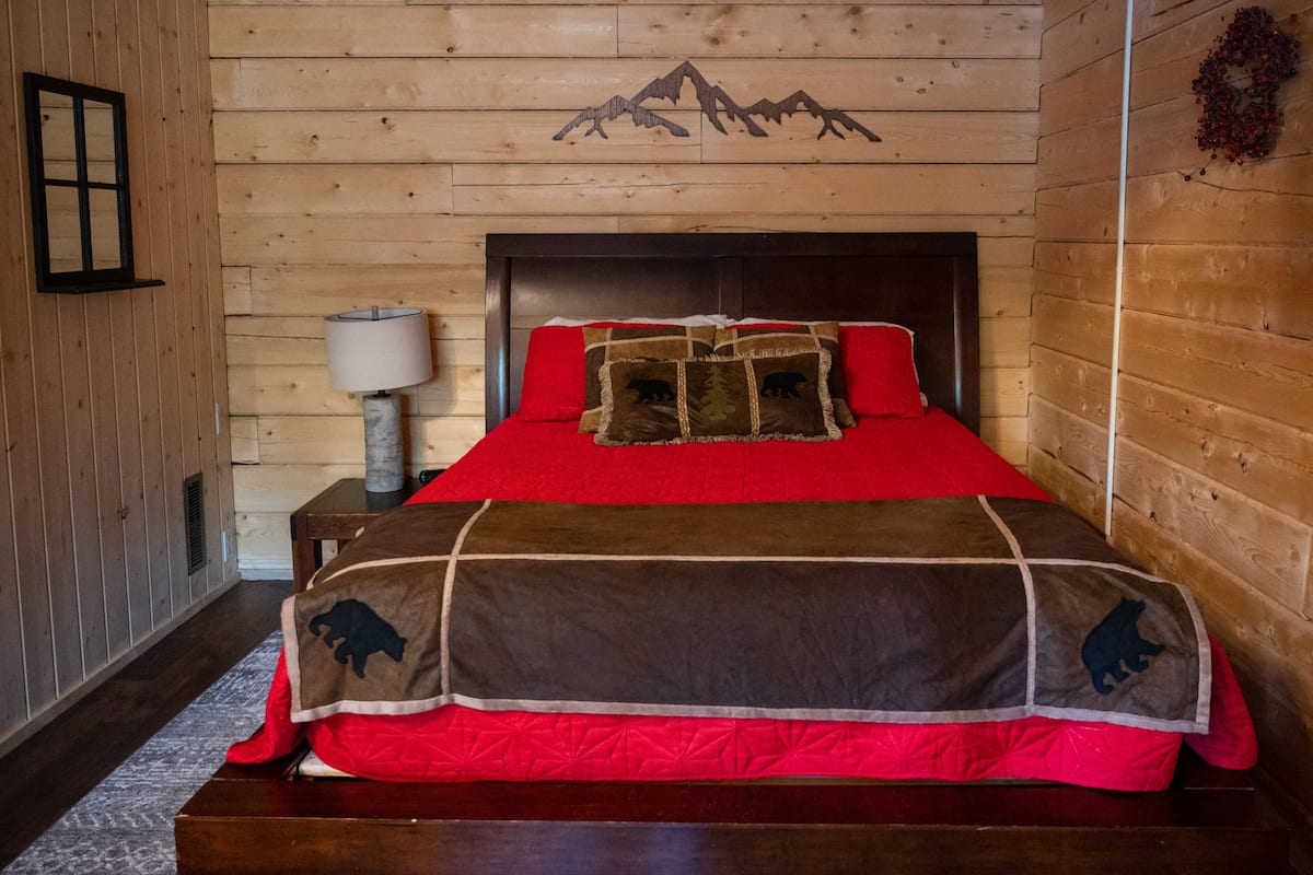 Una habitación con paneles de madera, una cama, una mesita de noche, una lámpara y un cuadro colgado en la pared de montañas.
