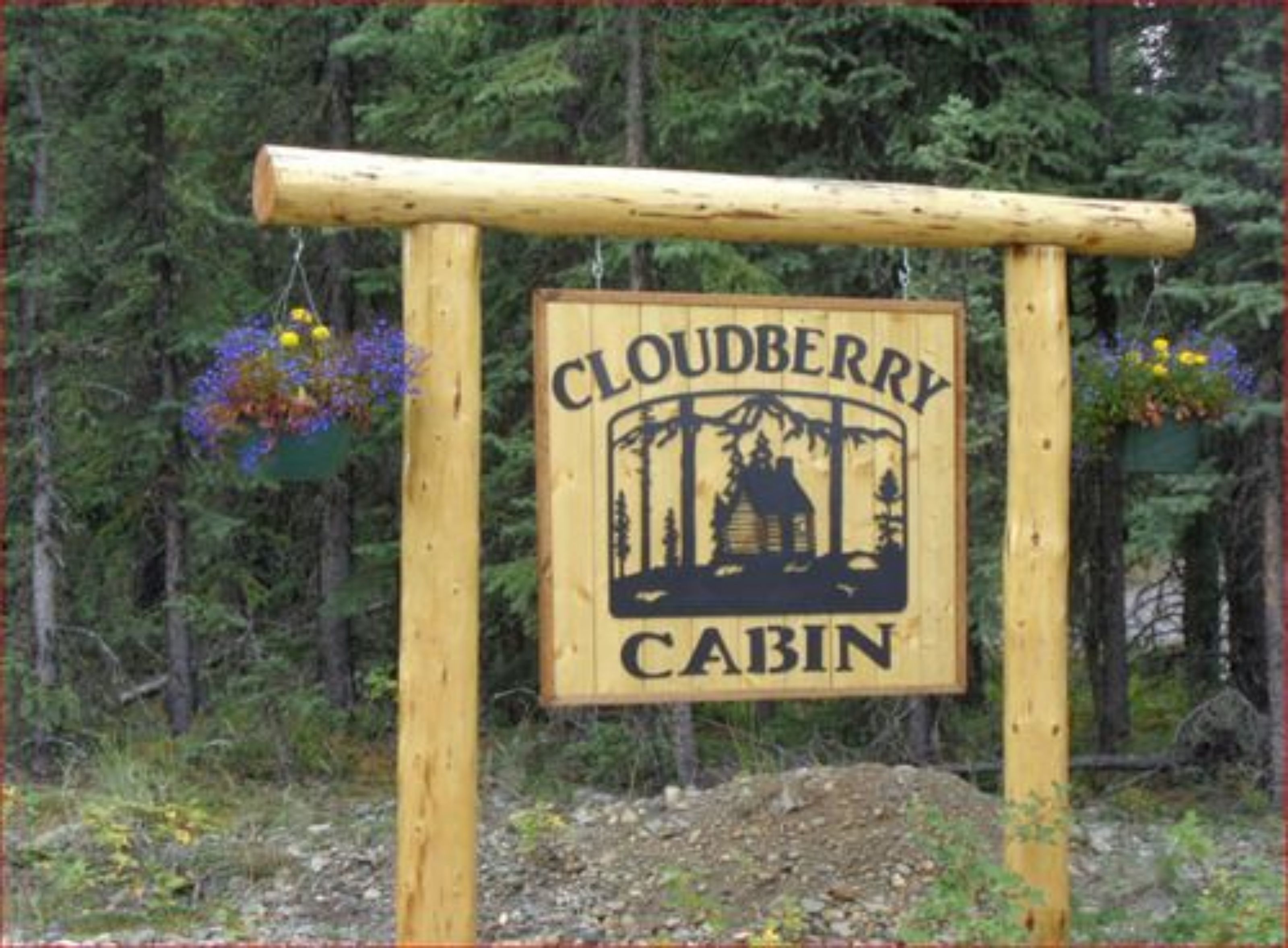 Puinen kyltti, jossa lukee ”Cloudberry Cabin”, ja jossa on mökin kuva sekä roikkuvia kukkaruukkuja.