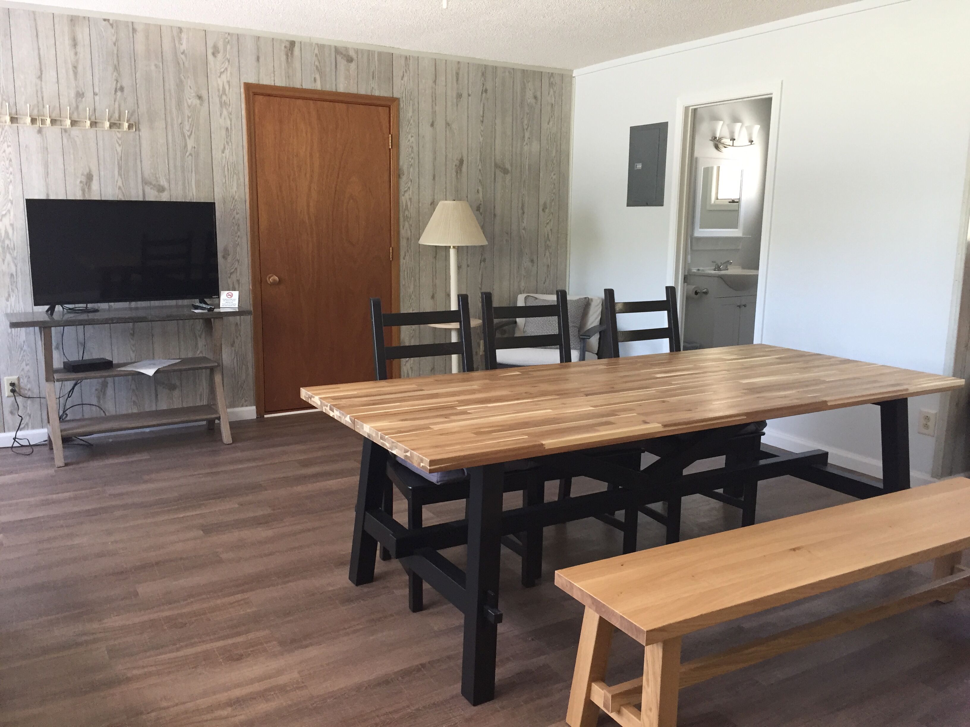 Un área de comedor con una mesa y sillas de madera, un banco, un televisor sobre un soporte y una lámpara.