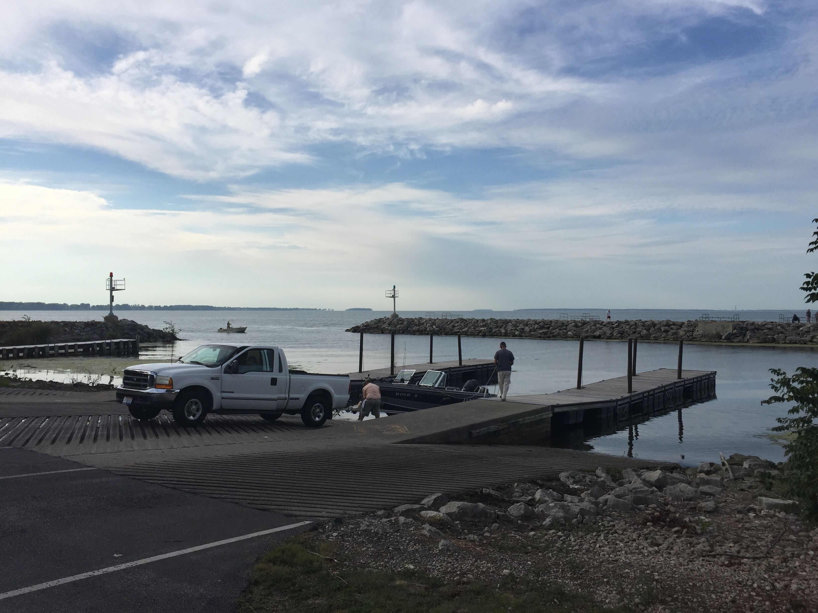 A dock with a truck parked nearby, a boat on the water, and a rocky shoreline.