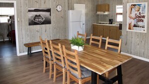 Un coin repas avec une table et des chaises en bois, un ventilateur de plafond, une horloge, et une cuisine en arrière-plan.