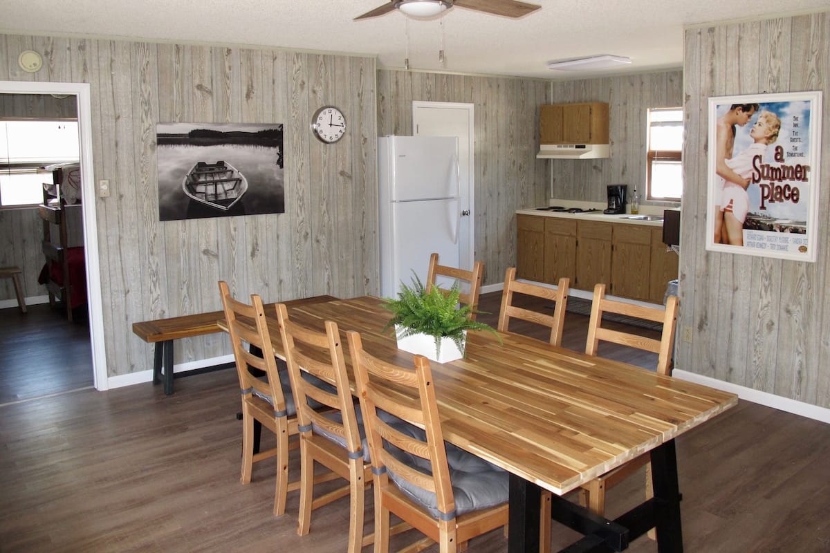 Un comedor con una mesa y sillas de madera, un ventilador de techo, un reloj y una cocina al fondo.