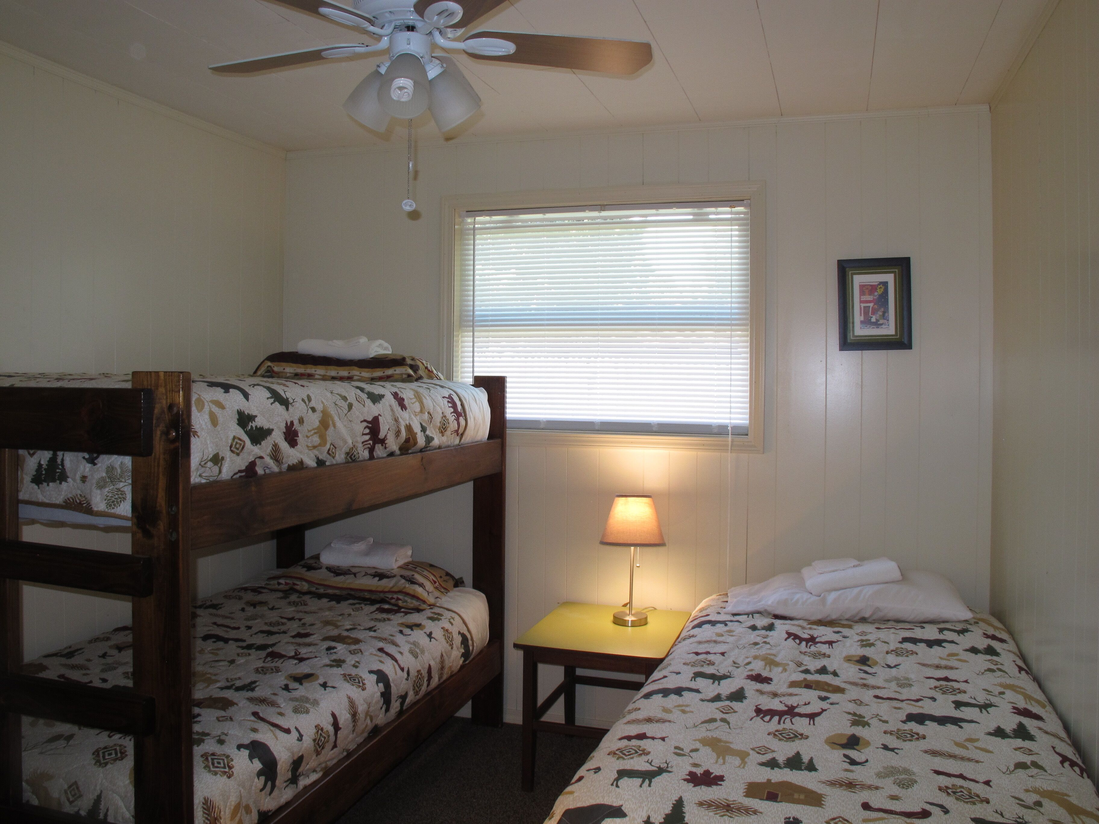 A dormitory room with two bunk beds, a ceiling fan, a window with blinds, a small table with a lamp, and a framed picture on the wall.