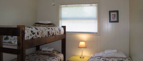 A dormitory room with two bunk beds, a ceiling fan, a window with blinds, a small table with a lamp, and a framed picture on the wall.