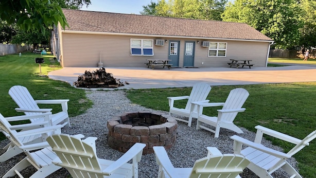 Un jardin avec un foyer entouré de chaises Adirondack blanches, une terrasse en bois et une maison avec un toit brun.