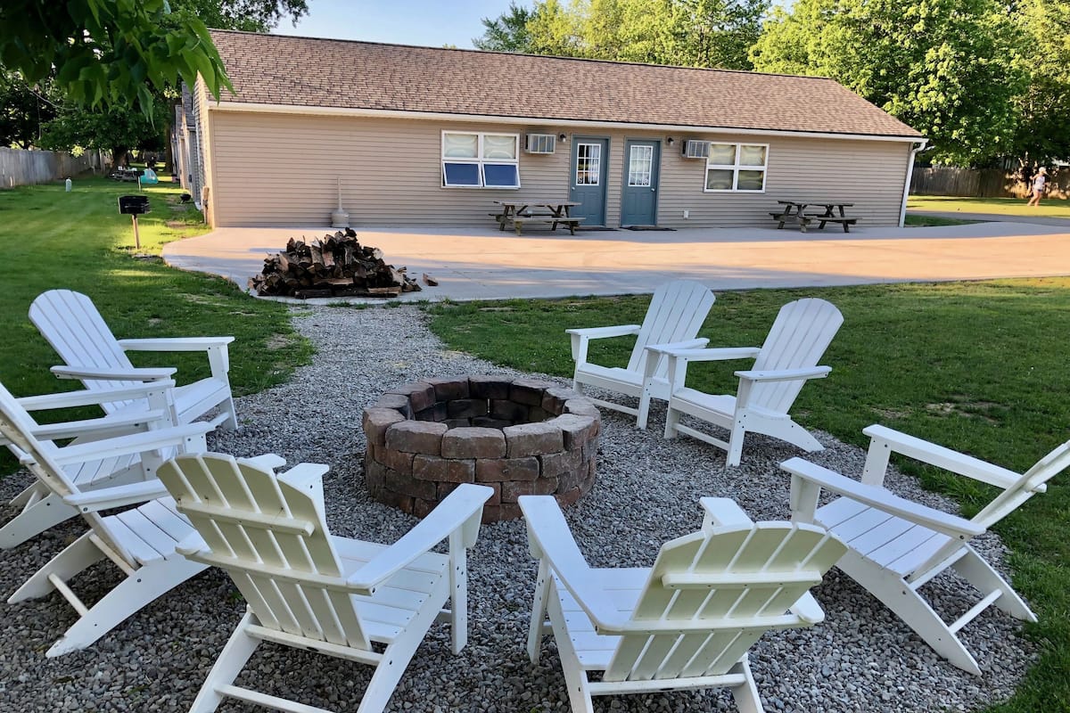 Patio con un brasero rodeado de sillas Adirondack blancas, una terraza de madera y una casa con techo marrón.