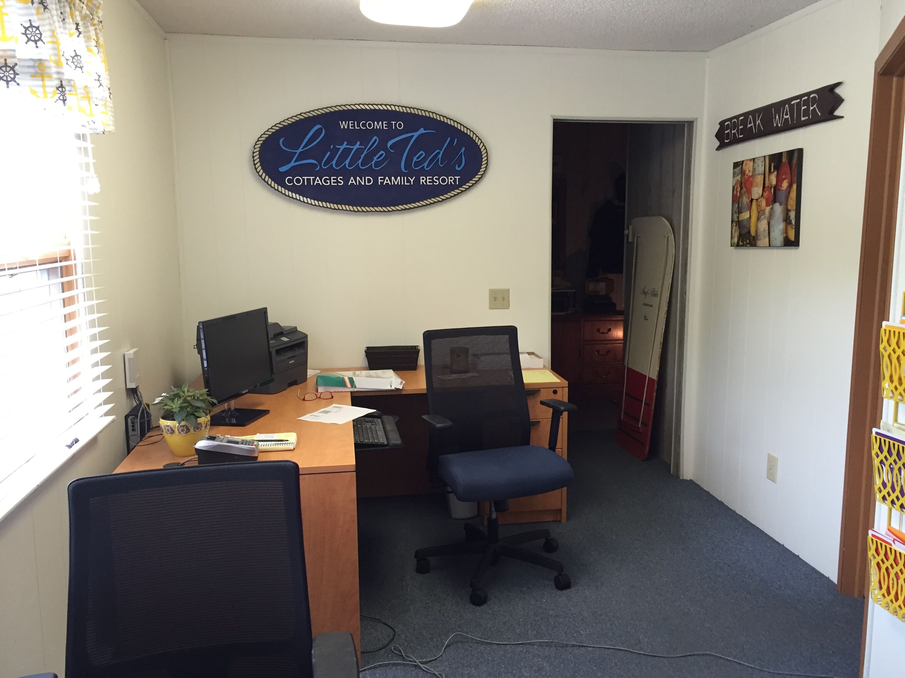 A home office with a desk, computer, and office chair. A sign on the wall reads "Welcome to Little Ted's Cottages and Family Resort."