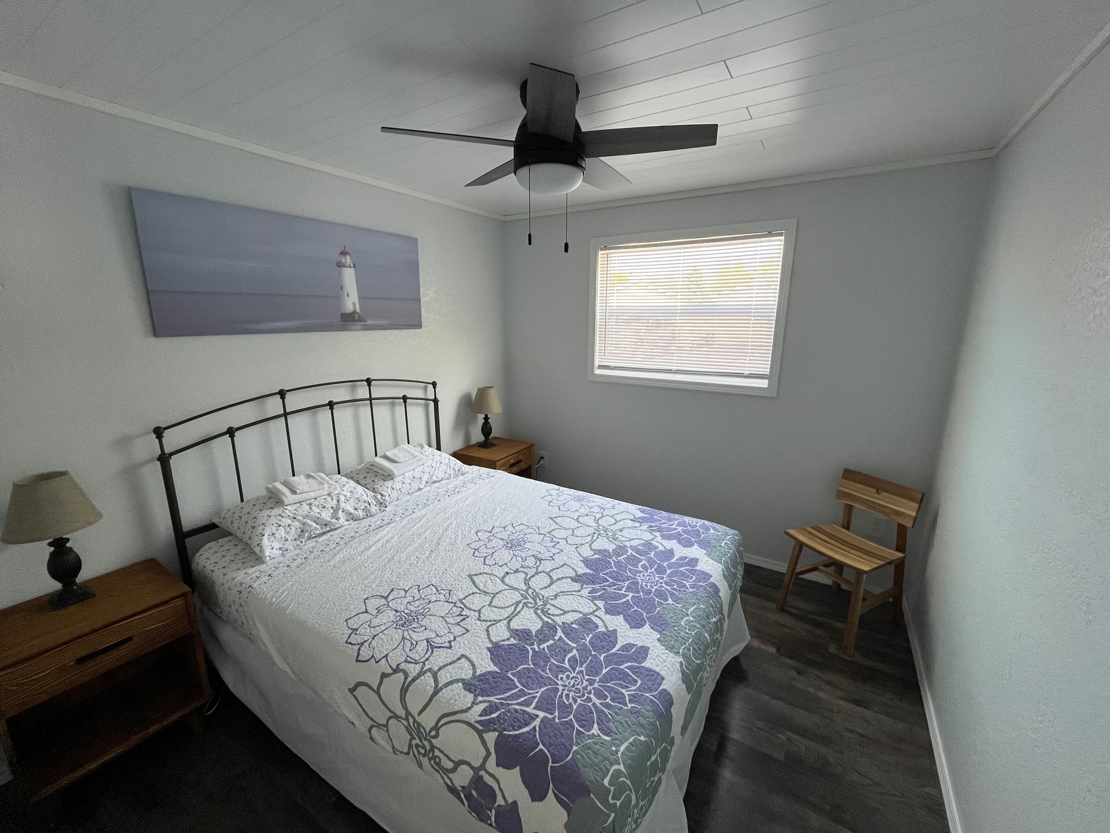 A bedroom with a bed, a ceiling fan, a nightstand with a lamp, a bench, a window with blinds, and a painting on the wall.