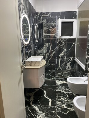 A bathroom with black marble tiles, a white sink, a white toilet, and a mirror with a light. - Hotel Red Apple (Mbrostar Ura)