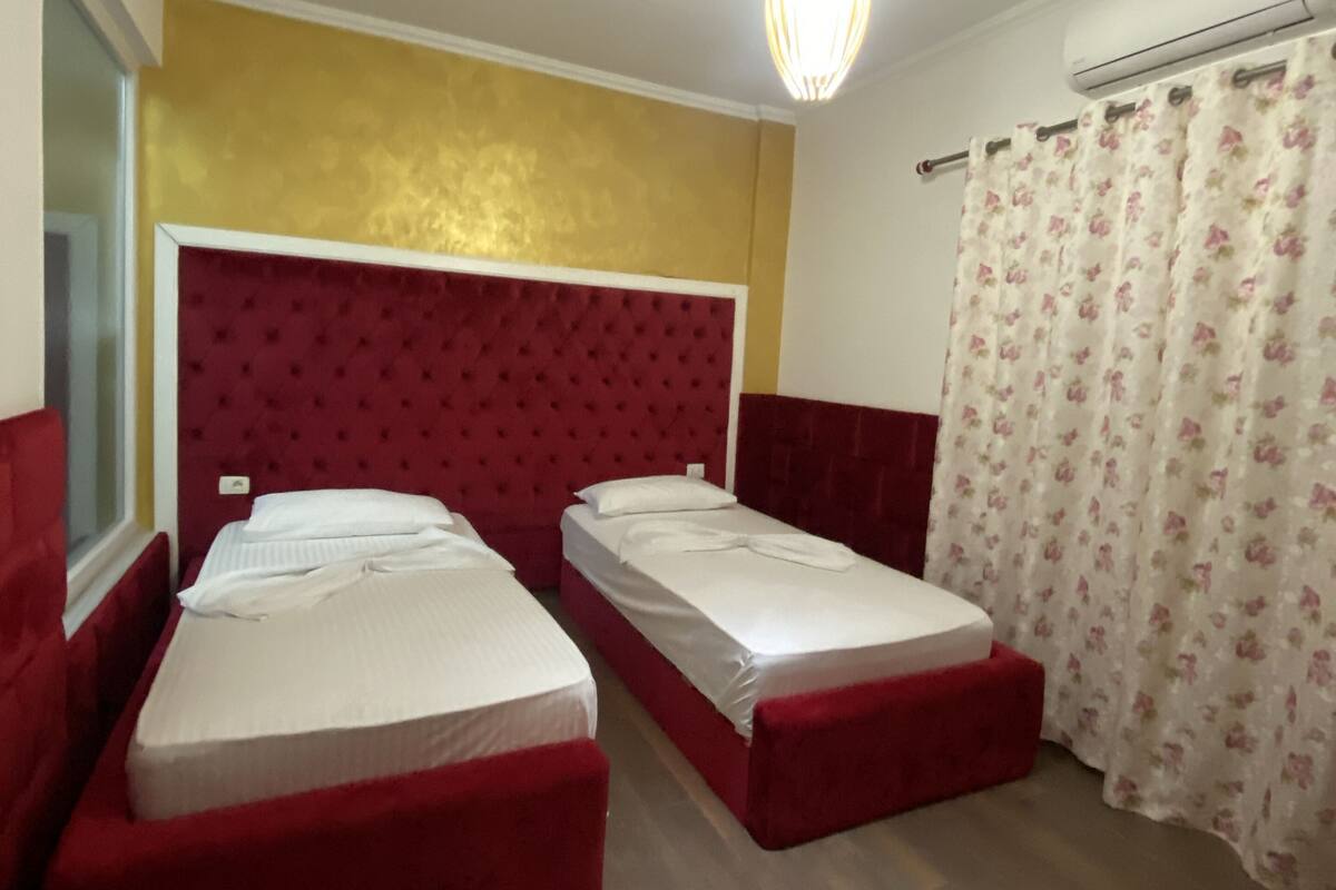 A hotel room with two beds, a red headboard, a window, and a floral curtain.