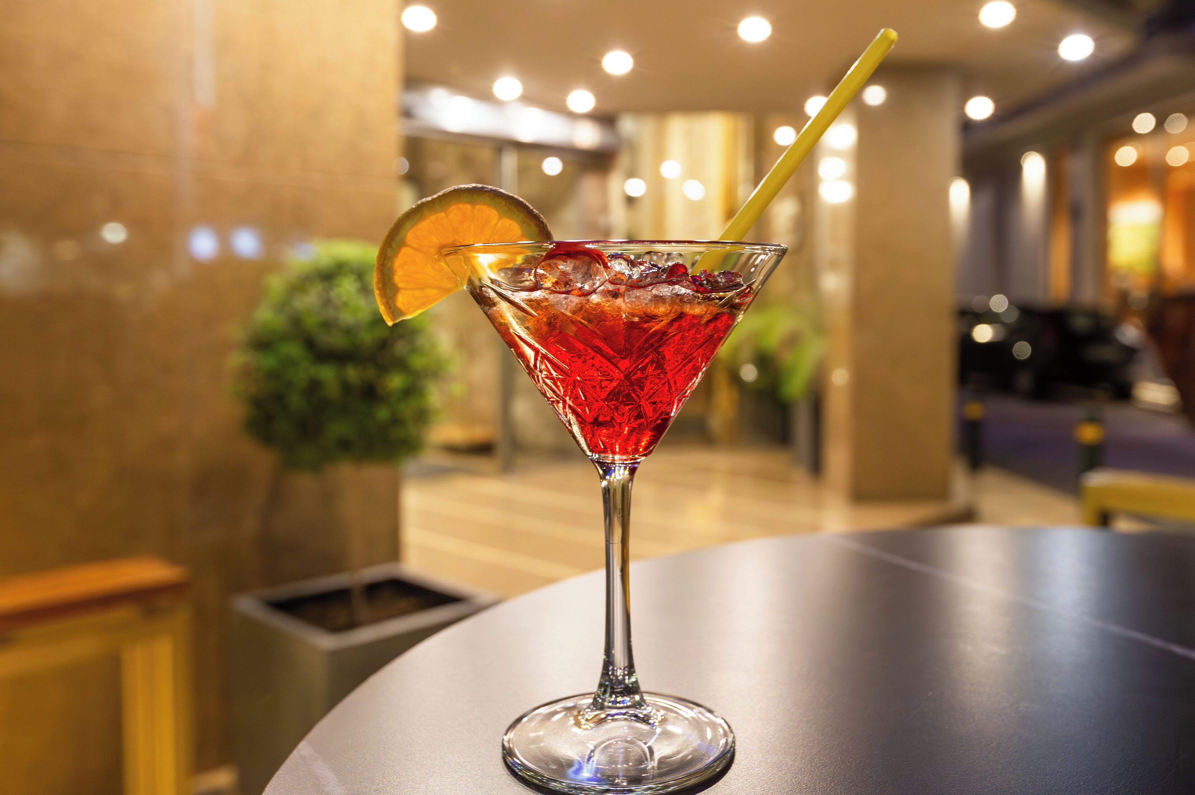 A martini glass with a red drink, ice, and a lemon slice on a table in a well-lit indoor setting.