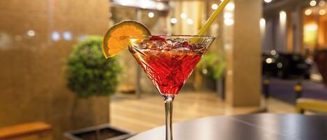 A martini glass with a red drink, ice, and a lemon slice on a table in a well-lit indoor setting.
