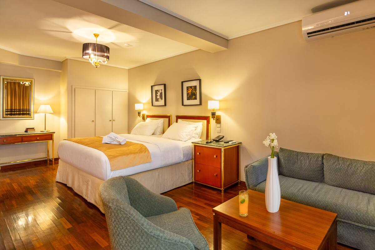 A hotel room with a bed, a desk, a chair, a sofa, a table with a vase and a drink, and wall-mounted lamps.