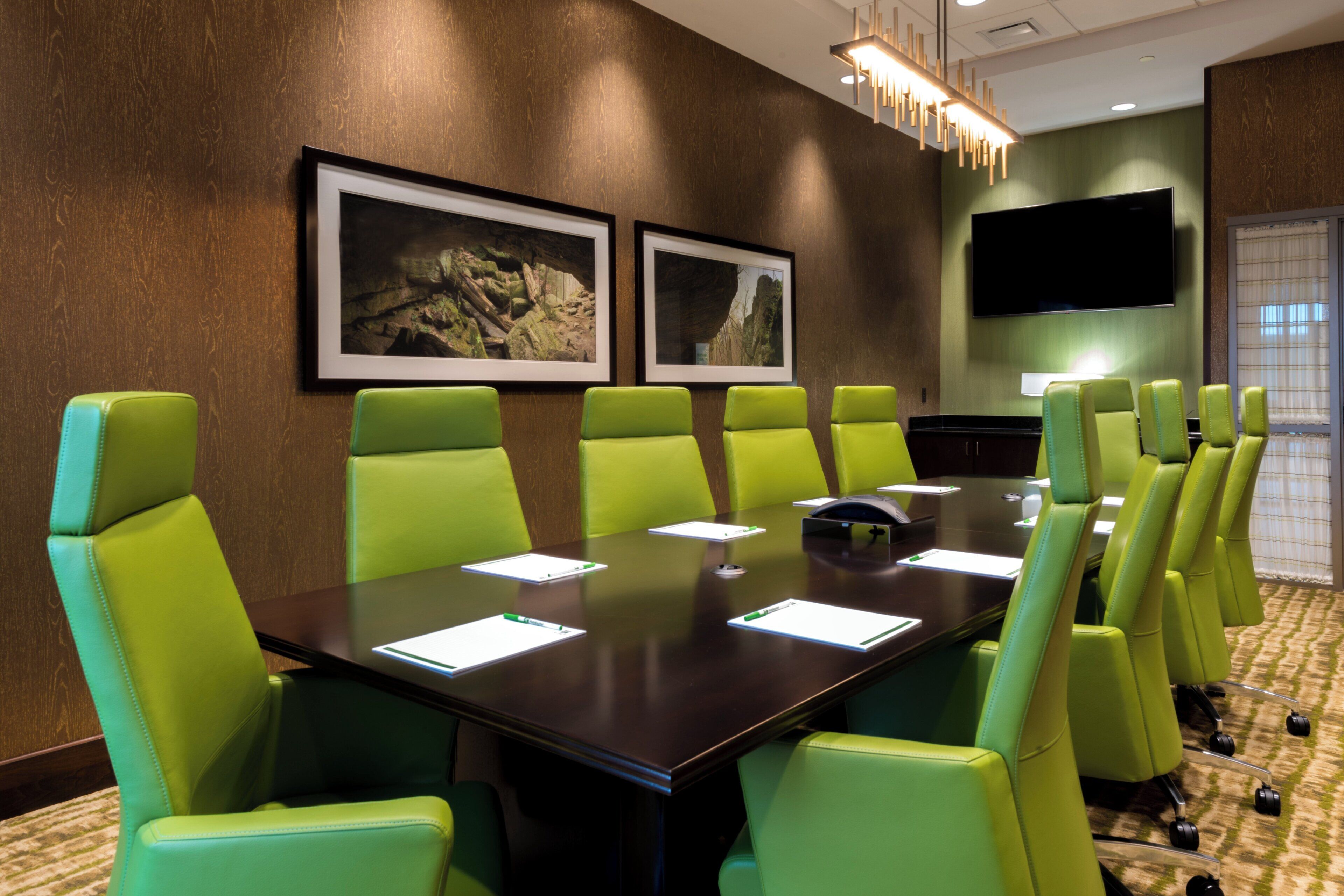 A conference room with a long table and green chairs, framed pictures on the wall, and a flat-screen TV.