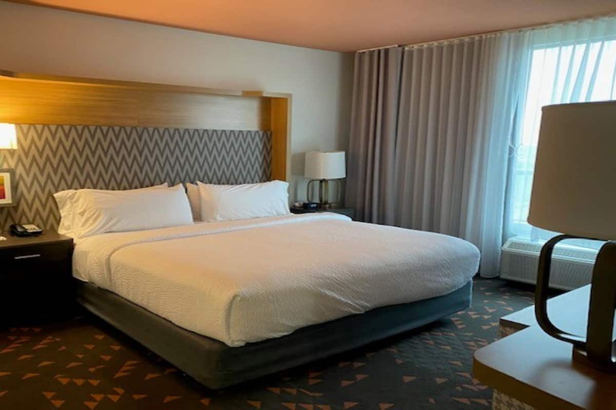 A hotel room with a large bed, a nightstand, a desk, a chair, a lamp, and a window with curtains.